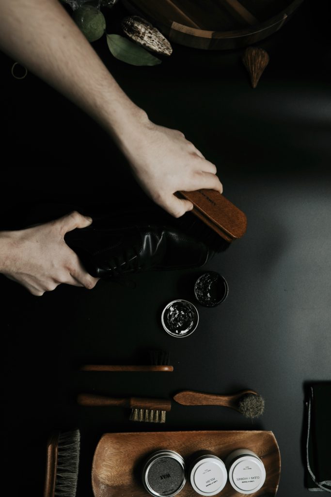 ブラシやクリーナーなどの手入れ道具が並べられた作業台で、職人が革靴を丁寧に手入れしている手元のクローズアップ。